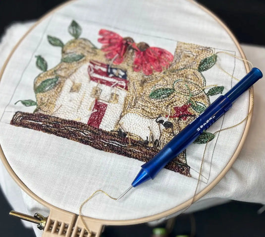 A punch needle embroidery project in progress, featuring a house design with a blue punch needle tool and thread.